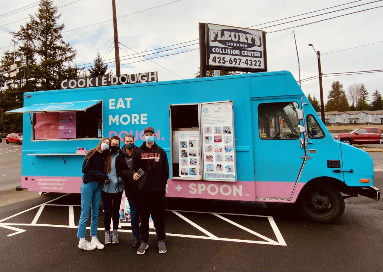 Three people at a blue cookie-dough food truck parked outside the shop.