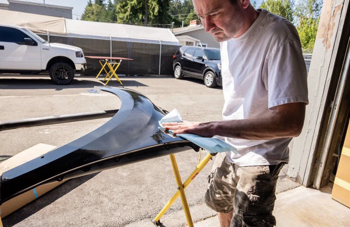 Technician sanding/preparing detached body panel near bay entrance.